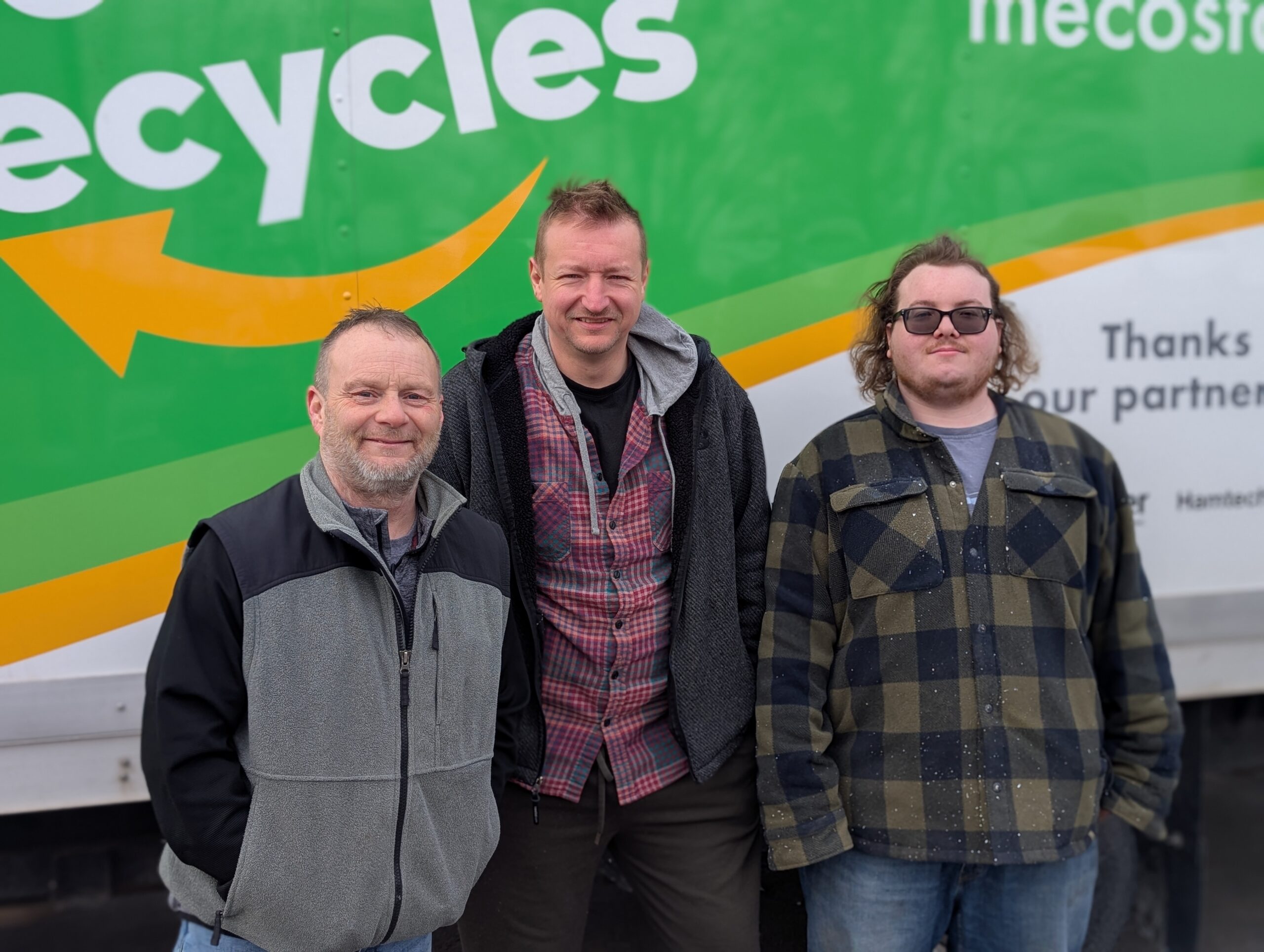 Three men stand in line posing for a picture. They are the staff members who pick up recycling from businesses. 